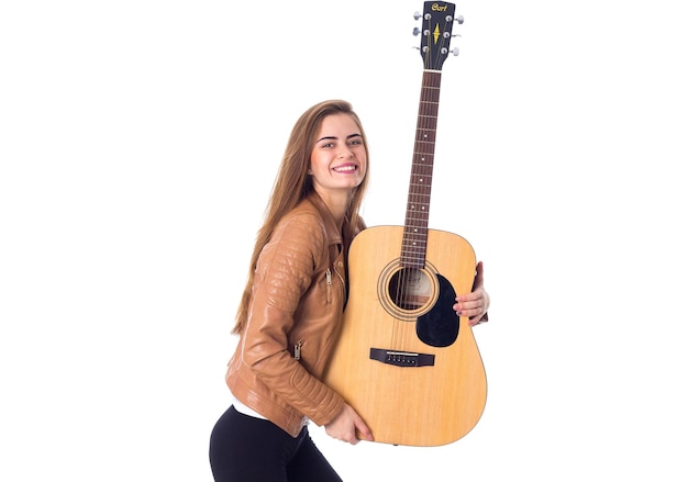 Encantadora joven de pelo largo con chaqueta marrón y pantalón negro sosteniendo una guitarra con fondo blanco en el estudio