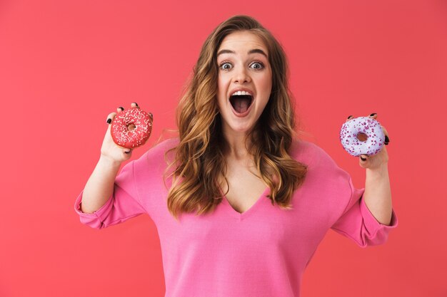 Foto encantadora joven rubia que se encuentran aisladas sobre pared rosa, sosteniendo donas