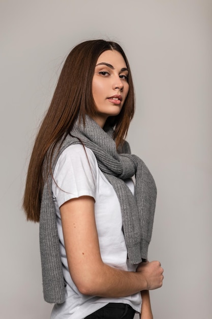 Encantadora jovencita linda con cabello moreno en ropa cómoda de moda con suéter de punto y camiseta blanca en el estudio. Hermosa mujer sobre fondo gris