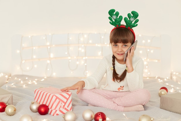 Encantadora linda niña de cabello oscuro con coletas sosteniendo el teléfono móvil en las manos, hablando por teléfono con amigos o padres, mirando a cámara, sentado en la cama con adornos navideños.