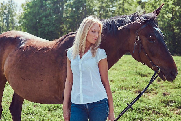 Encantadora loira linda vestindo uma blusa branca e jeans em pé com um cavalo em uma zona rural.