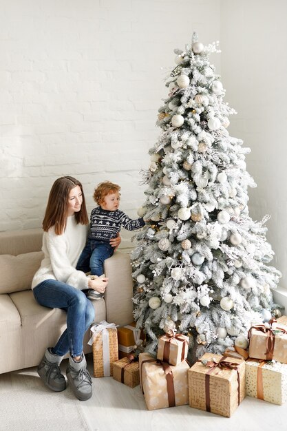 Encantadora madre en el suéter blanco sosteniendo a su pequeño hijo cerca del árbol de Navidad y cajas de regalo en la sala de estar de la casa