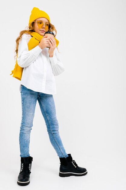 encantadora menina adolescente elegante e sorridente com um chapéu de inverno com um copo de papel em uma parede branca