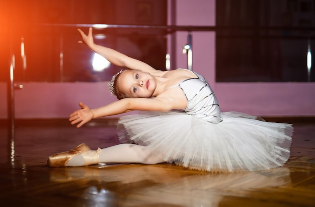 Encantadora menina bailarina de tutu branco realizando poses de balé no chão no fundo do estúdio Uma pequena bailarina doce sentada no chão de madeira em um estúdio de dança
