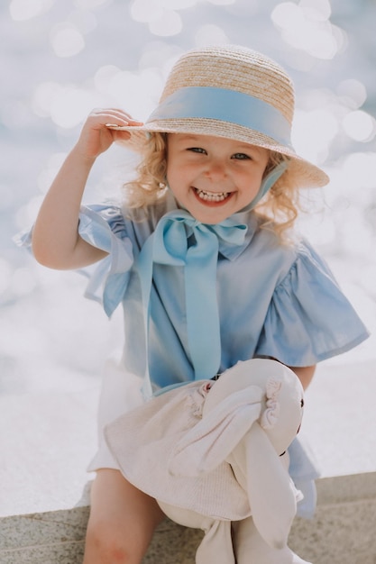 encantadora menina loira com um chapéu de palha e um vestido azul brinca com um coelho de pelúcia ao ar livre