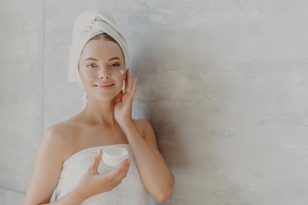 Encantadora modelo aplica crema hidratante sostiene tarro de crema rutina de belleza poses envueltas en una toalla contra la pared gris