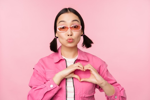 Encantadora modelo coreana con gafas de sol de moda muestra un signo de cuidado del corazón Te amo gesto sobre fondo rosa