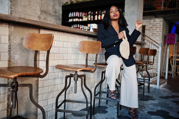 Encantadora modelo de mulher afro-americana em jaqueta preta e bolsa de cintura relaxando no café durante o tempo livre