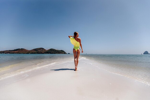 Encantadora modelo delgada en un traje amarillo con una hermosa figura posando en la arena blanca junto al mar azul, sosteniendo su mano cerca de su cabello. Vista trasera