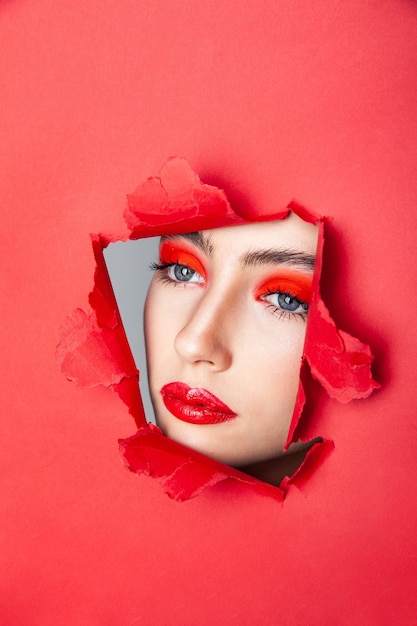 Encantadora modelo femenina con maquillaje rojo mirando a otro lado a través de papel rasgado en estudio