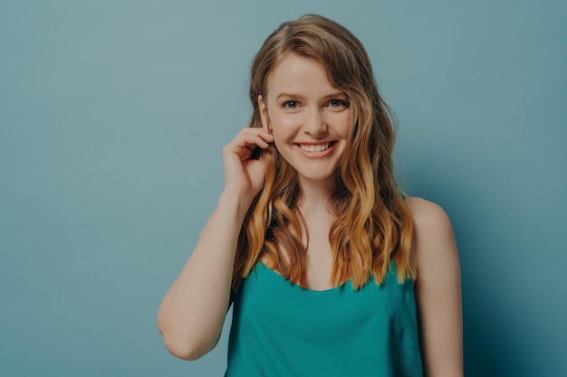 Encantadora modelo femenina mirando a la cámara con una sonrisa radiante metiéndose el pelo ondulado detrás de la oreja