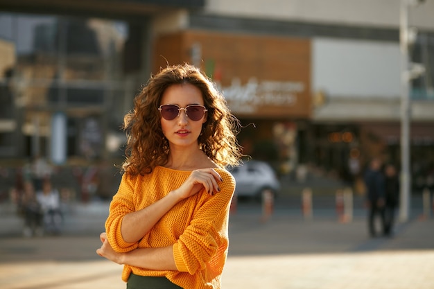 Encantadora modelo joven con pelo rizado en gafas de sol con ropa de moda y caminando en la calle soleada. Espacio para texto