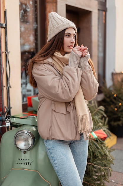 Foto encantadora modelo de mujer joven con un sombrero de punto de moda, suéter y una elegante chaqueta está de pie cerca de un scooter verde vintage en la ciudad. vacaciones y estilo de invierno femenino.