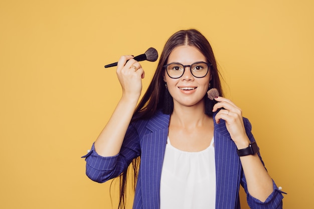 Encantadora morena en gafas con el pelo largo vestido con traje azul oscuro linda sonrisa, se maquilla durante la cuarentena, sobre fondo amarillo con espacio de copia.