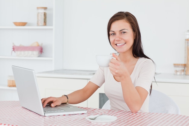 Encantadora morena usando su computadora portátil y tomando un té