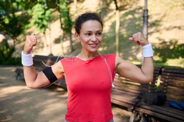 Encantadora mujer activa confiada deportista apretando los puños tensando el brazo y los músculos bíceps sonriendo de pie en el parque de la ciudad durante el entrenamiento deportivo