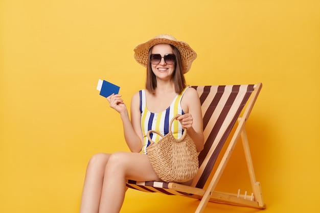 Encantadora mujer adulta optimista con traje de baño a rayas y sombrero con pasaporte y boletos para viajar al sur del país en el resort en verano