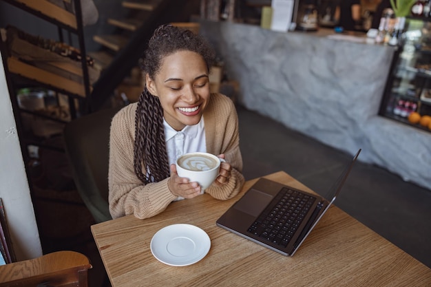 Encantadora mujer afroamericana trabajando remotamente en una laptop y disfrutando de un descanso para tomar café en un acogedor café