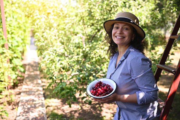 Encantadora mujer agricultora ecológica sonriendo con dientes mirando a la cámara sosteniendo un tazón con cerezas recién cosechadas