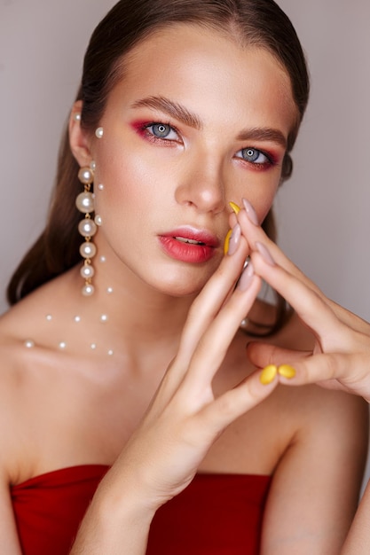 Encantadora mujer con aretes de perlas y vestido rojo mirando a la cámara con fondo gris