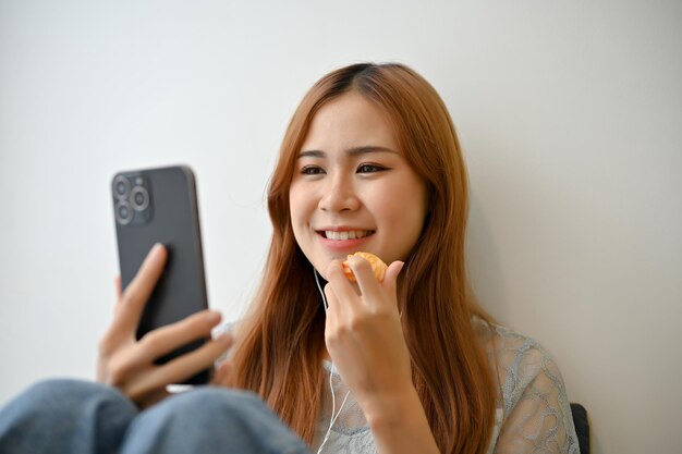 Encantadora mujer asiática bastante joven comiendo una galleta y usando un teléfono inteligente en la sala de estar