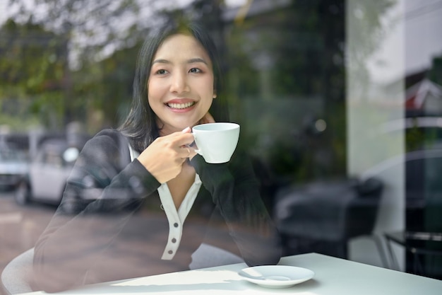 Encantadora mujer asiática bebiendo café matutino en la cafetería mirando por la ventana