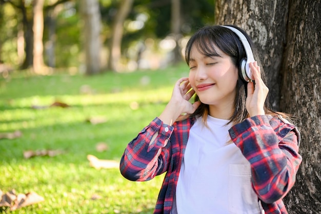 Encantadora mujer asiática escuchando música a través de sus auriculares mientras se relaja sentada bajo el árbol