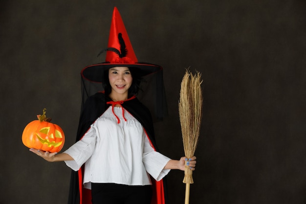 Encantadora mujer asiática joven sonriente en traje de bruja con calabaza de Halloween y escoba mirando a la cámara contra el fondo gris oscuro