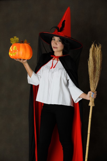 Encantadora mujer asiática joven sonriente en traje de bruja con calabaza de Halloween y escoba mirando a la cámara contra el fondo gris oscuro