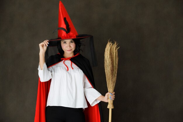 Encantadora mujer asiática joven sonriente en traje de bruja con sombrero de Halloween y escoba mirando a la cámara contra el fondo gris oscuro