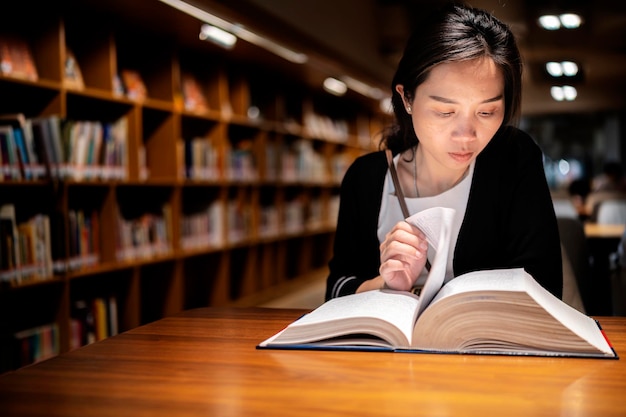 Una encantadora mujer asiática lee un libro en la biblioteca de la universidad para relajarse y educarse