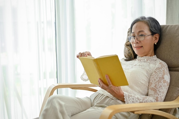 Una encantadora mujer asiática de mediana edad con anteojos se relaja leyendo un libro en su acogedor sillón