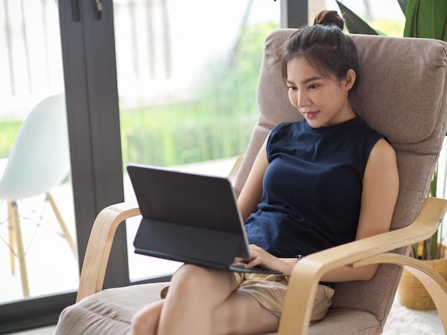 Encantadora mujer asiática relajándose en casa sentada en un sillón y viendo películas en una tableta digital