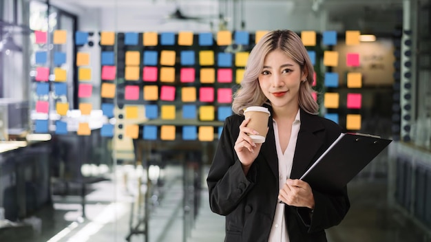 Encantadora mujer asiática con una sonrisa de pie sosteniendo papeles y una taza de café en la oficina