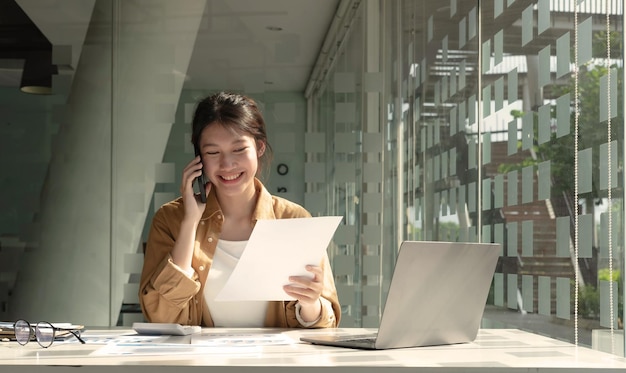 Encantadora mujer asiática con una sonrisa de pie sosteniendo papeles y teléfono móvil en la oficinaxAEncantadora mujer asiática con una sonrisa de pie sosteniendo papeles y teléfono móvil en la oficinaxA