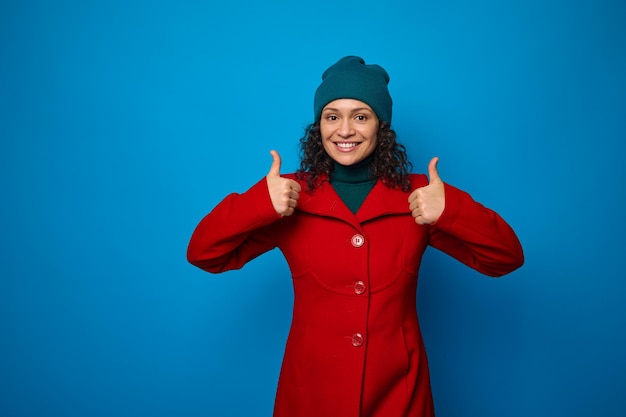 Encantadora mujer bonita atractiva maravillosa en abrigo rojo brillante y sombrero verde sonriendo con alegre sonrisa dentuda, mirando a cámara y mostrando los pulgares hacia arriba, posando sobre fondo azul con espacio de copia