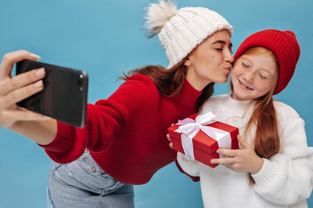 Encantadora mujer de cabello moreno con sombrero blanco de invierno, falda de mezclilla y camisa roja besa a su hermana menor con un atuendo fresco y ligero