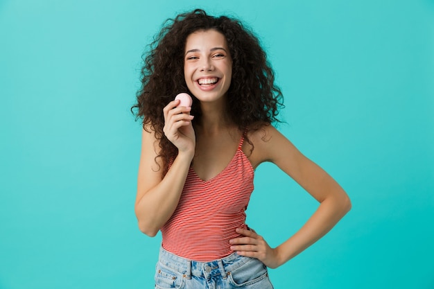 Encantadora mujer con cabello rizado sonriendo y comiendo galletas macaron, aislado sobre pared azul