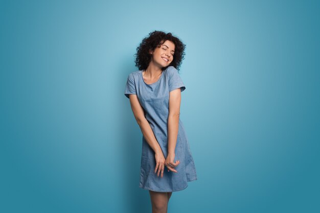 Encantadora mujer caucásica con cabello rizado con un vestido azul de verano está sonriendo en una pared