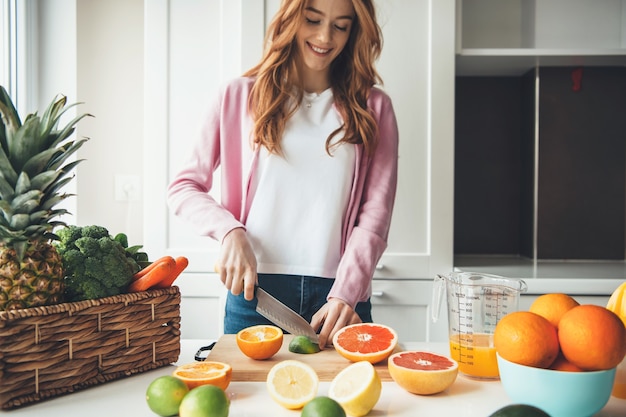 Encantadora mujer caucásica con pelo rojo y pecas está cortando frutas con un cuchillo y haciendo jugo en casa