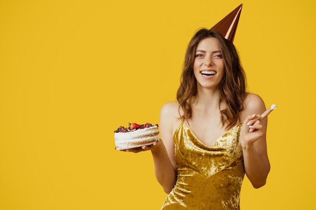 Encantadora mujer caucásica en vestido y sombrero de cumpleaños degustando un delicioso pastel de cumpleaños en el espacio de copia de fondo amarillo