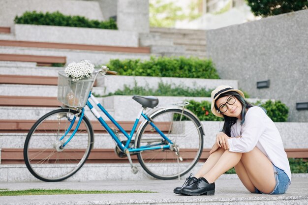 Encantadora mujer chiense joven sonriente en pantalones cortos de mezclilla sentada en el suelo ext a su bicicleta y mirando a la cámara