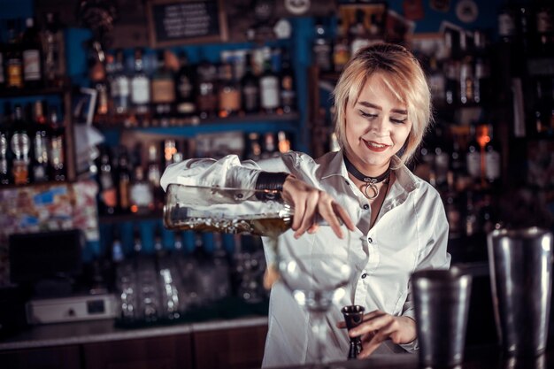 Encantadora mujer coctelería formula un cóctel mientras está de pie cerca del mostrador del bar en el pub