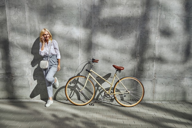 Encantadora mujer hablando por teléfono móvil con amigos mientras está de pie cerca de la bicicleta aislada en la pared gris al aire libre