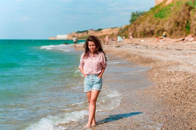 Una encantadora mujer joven con cabello largo y vistiendo una camiseta camina por la playa en un día soleado