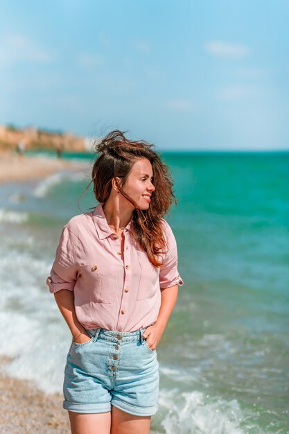 Una encantadora mujer joven con cabello largo y vistiendo una camiseta camina por la playa en un día soleado