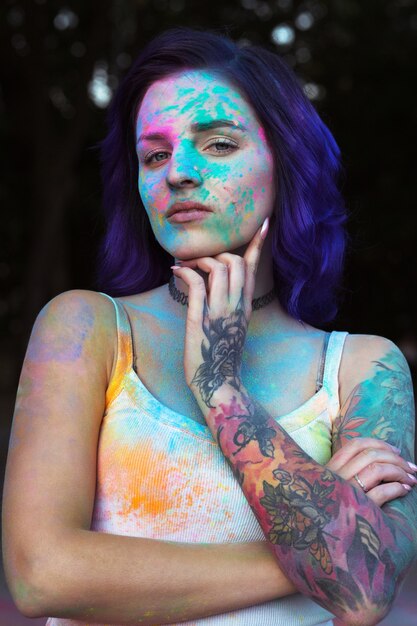 Encantadora mujer joven con cabello morado y tatuaje en su brazo cubierto con coloridos polvos Holi