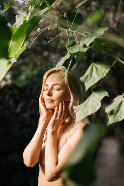 Encantadora mujer joven caucásica sensual posando en el bosque con los ojos cerrados, disfrutando del sol y la naturaleza del verano, tocando la cara, usando cosméticos orgánicos, aplicando protector solar. Cuidado de la piel, concepto de spa.