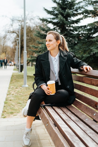 Encantadora mujer joven en elegante abrigo descansando en un banco durante el descanso para tomar café al aire libre Sonriente dama elegante sosteniendo una taza de café desechable y mirando hacia otro lado en el parque