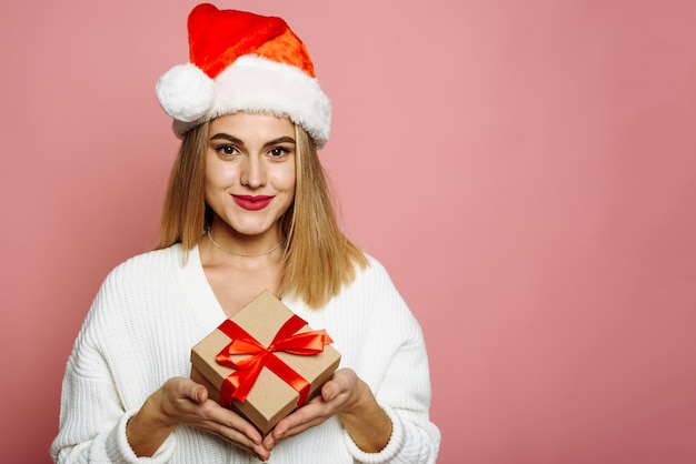 Encantadora mujer joven con gorro de Papá Noel está sosteniendo un regalo de Navidad sonriendo sobre un fondo rosa